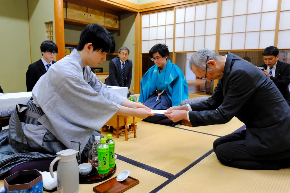 立会人の中田功八段（手前右）に封じ手を渡す藤井聡太王位（同左）。中央は伊藤匠二冠=26日午後6時18分、徳島市の「渭水苑」